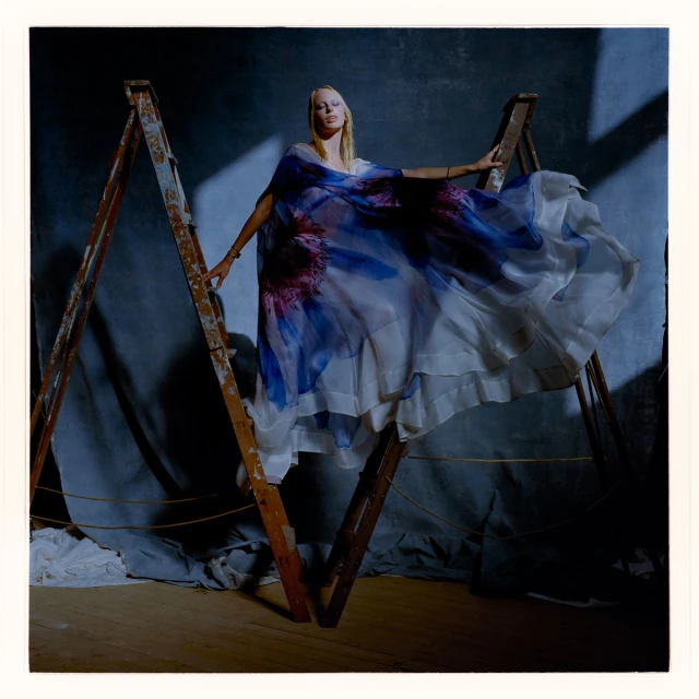 A blonde woman in a blue and white floral patterned dress poses on a weathered wooden A-frame ladder in a photo studio setting.