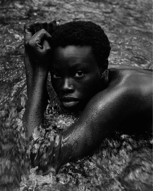 Close-up black and white portrait of a Black woman partially submerged in flowing water with her arms raised and hands near her face.