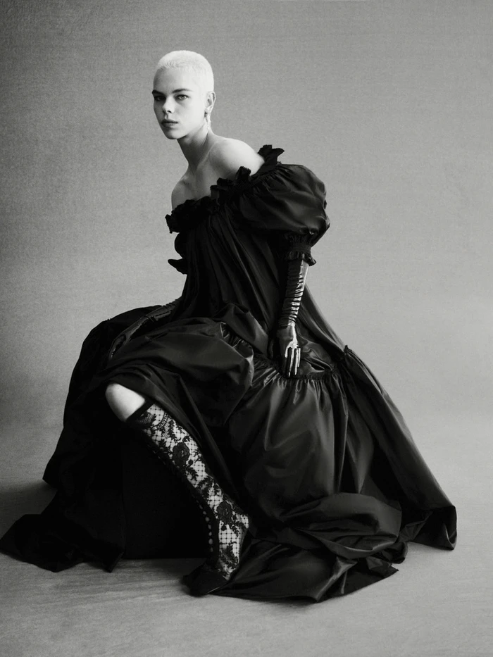 Model with a buzzed haircut seated in a voluminous dark off-the-shoulder dress with patterned stockings against a grey backdrop