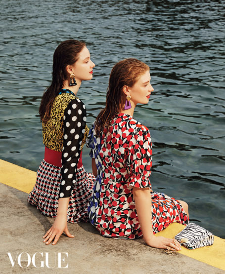 Two women sit by the water's edge, wearing vibrant patterned dresses and statement earrings, with a small purse beside them.