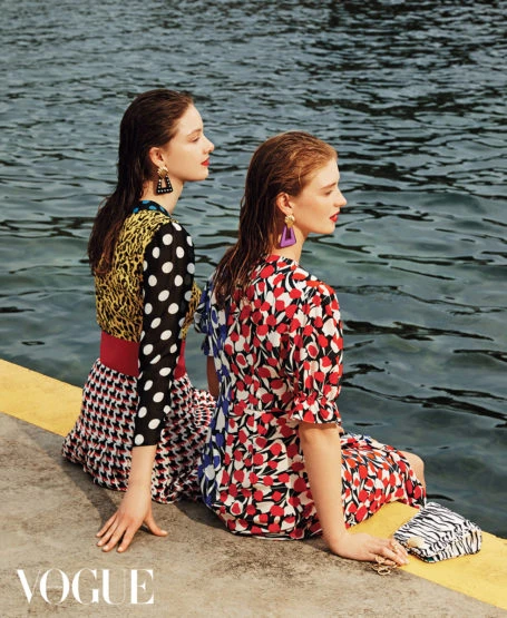 Two women sit by the water's edge, wearing vibrant patterned dresses and statement earrings, with a small purse beside them.