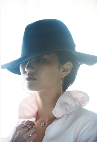 Portrait of a woman with a black fedora, large gold hoop earrings, and a pink ruffled collar against a bright white background.