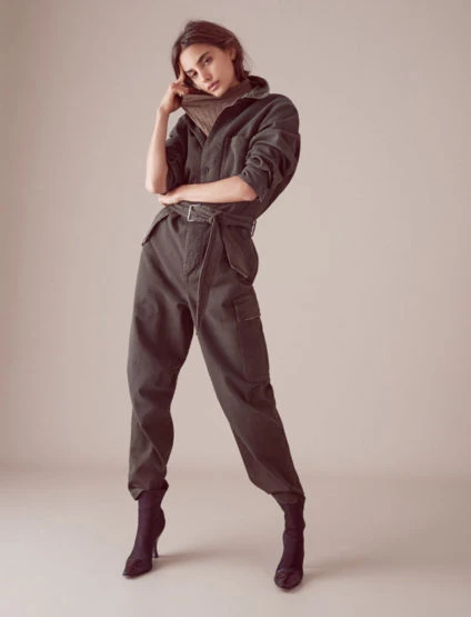 An adult woman poses in a green jumpsuit with a turtleneck sweater and heeled boots against a neutral backdrop in a studio shot.