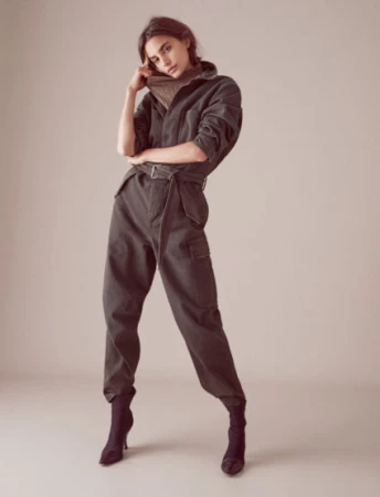 An adult woman poses in a green jumpsuit with a turtleneck sweater and heeled boots against a neutral backdrop in a studio shot.