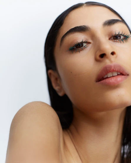 Close-up portrait of a young adult woman with green eyeliner and a fresh complexion, set against a bright white background.