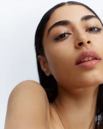 Close-up portrait of a young adult woman with green eyeliner and a fresh complexion, set against a bright white background.