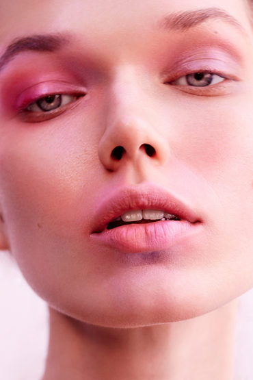 Close-up portrait of a woman with vibrant pink and violet eyeshadow and flushed pink cheeks by make-up artist Rae Morris.