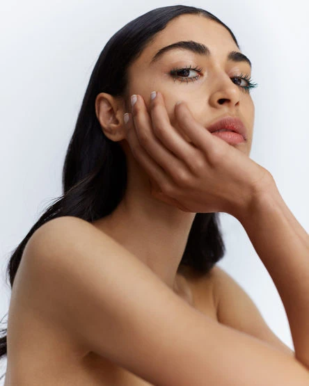 Close-up portrait of a dark-haired woman with flawless skin and hand near her face, posing in a neutral indoor environment.