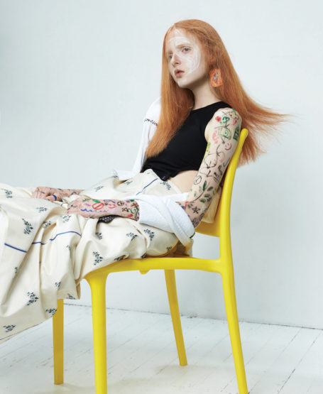 A pale-skinned woman with orange hair sits on a yellow chair, wearing a black top, patterned trousers, and earrings against a white background.