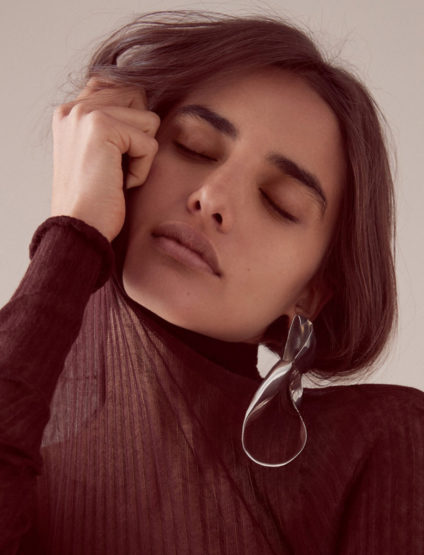 Portrait of a woman with dark hair and closed eyes wearing a sheer top and large earrings. Neutral background and soft lighting.