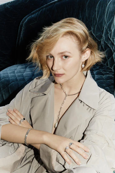 Blonde woman in beige trench coat poses indoors with jewelry. She has her arms crossed and is sitting in front of a plush blue couch.