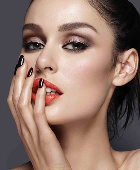Close-up studio shot of a woman with dark eye makeup, orange lipstick, and dark nail polish posing against a grey background.
