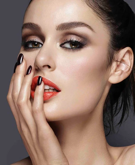 Close-up studio shot of a woman with dark eye makeup, orange lipstick, and dark nail polish posing against a grey background.