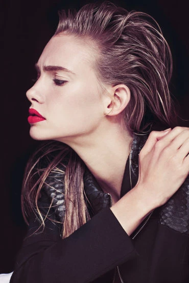 Portrait of a fashionable young woman with slicked-back wet hair, wearing a jacket, and bright red lipstick against a dark backdrop.