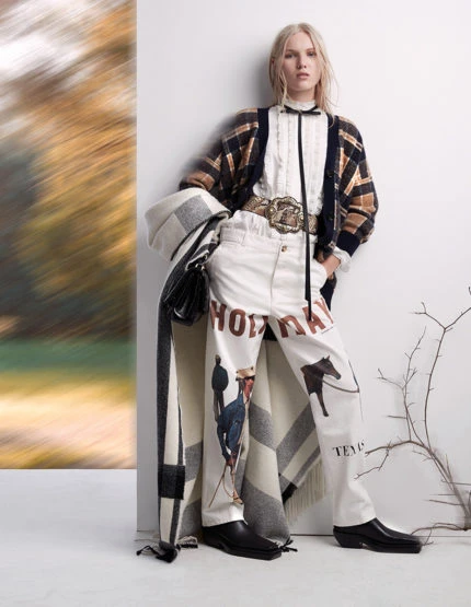 Fashion shot of a young, fair-skinned woman posing indoors in a jacket, white pants with horse and rider graphics, and a vintage belt.