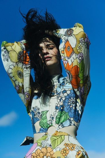 A young woman in a colorful floral-print dress stands against a blue sky, with her dark hair tousled around her face and shoulders.