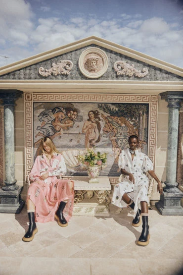 Two stylish women in printed dresses and black platform boots pose outdoors against a decorative mosaic wall under a sunny sky.