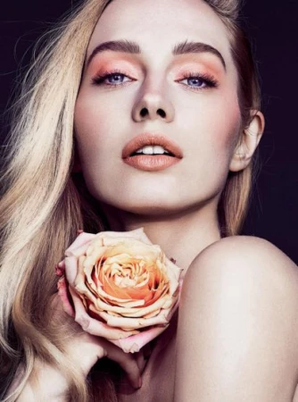 Close-up beauty portrait of a blonde woman holding a peach-toned rose with peach eye shadow and neutral lipstick.