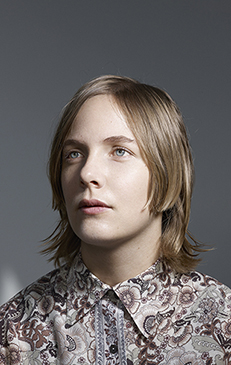 Portrait of a young adult with light blonde hair and wearing a patterned collared shirt, looking upwards against a gray backdrop.