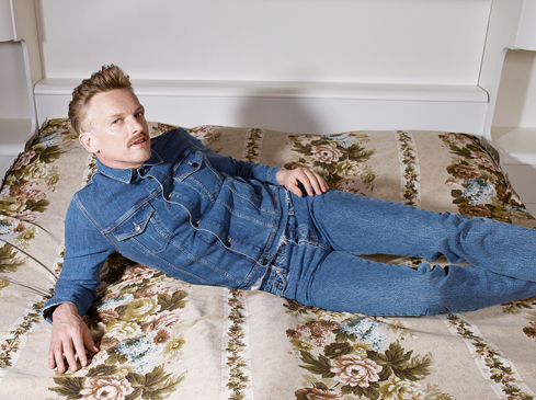 Man in denim jacket and jeans lies on floral bedspread, posing against a white headboard in an indoor setting under soft lighting.