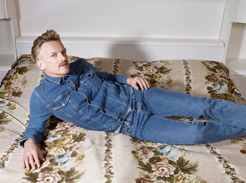 Man in denim jacket and jeans lies on floral bedspread, posing against a white headboard in an indoor setting under soft lighting.
