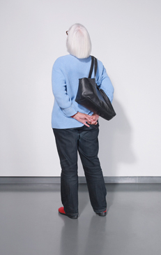 Back view of an elderly woman with short white hair wearing a blue sweater, dark pants, and red shoes, carrying a black bag.