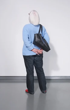 Back view of an elderly woman with short white hair wearing a blue sweater, dark pants, and red shoes, carrying a black bag.