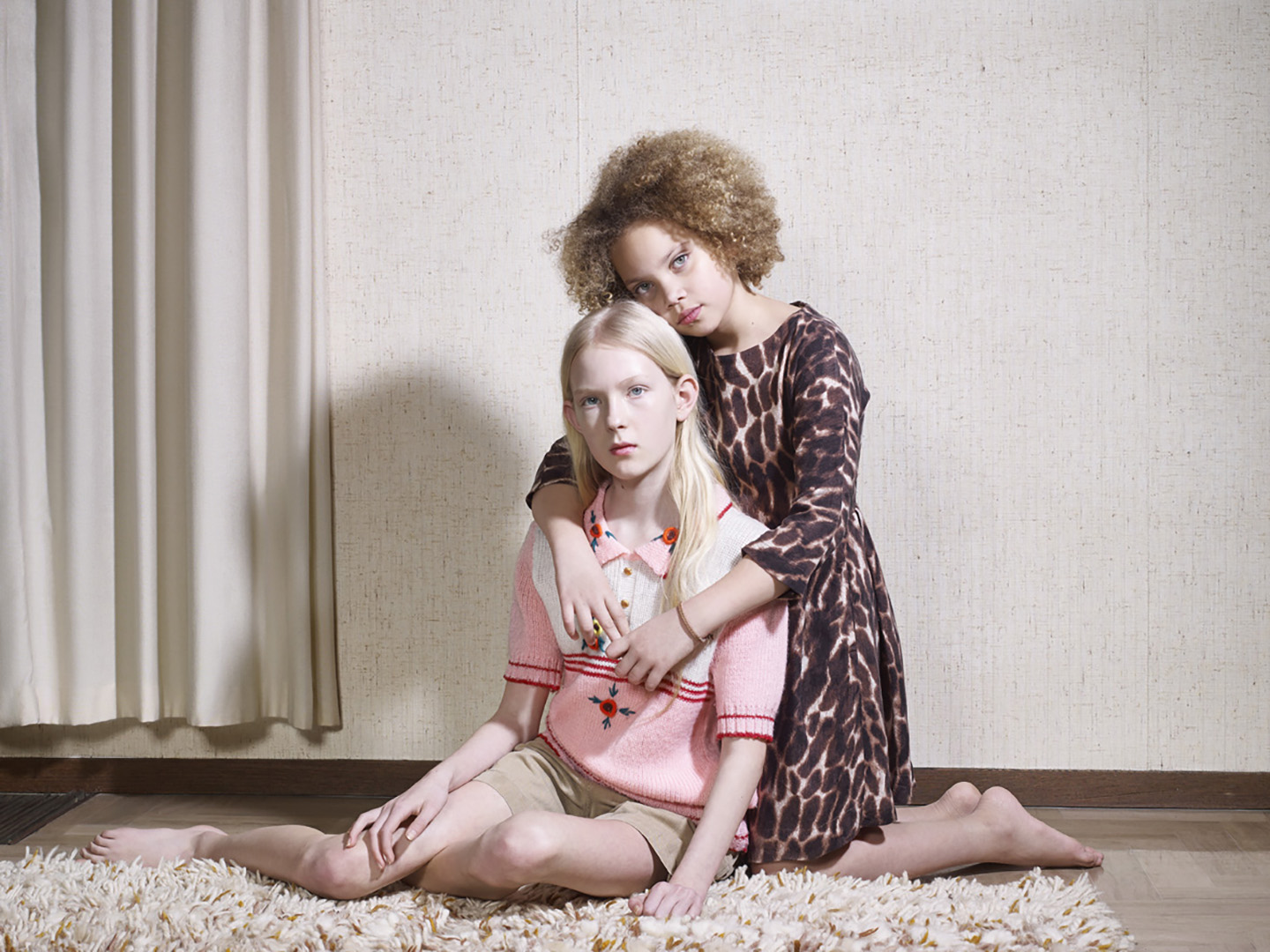 Two young girls pose indoors; one with blonde hair seated on a rug, embraced by another with curly hair wearing a leopard-print dress.