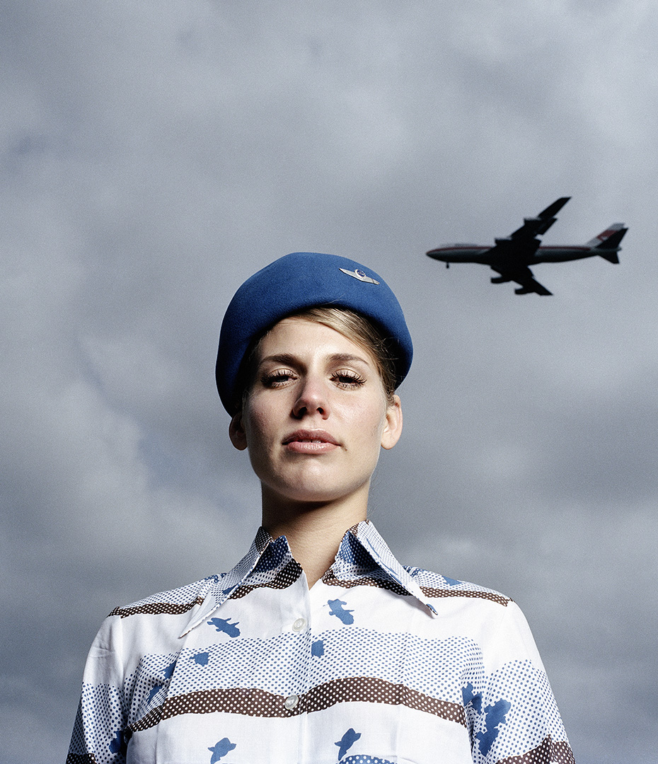 Attractive woman in uniform with blue hat and patterned blouse, airplane in background, low angle. Aviation theme. Cloudy sky.