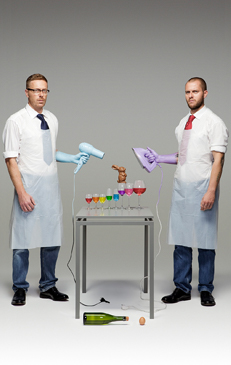 Two adult males wearing aprons with objects arranged on a table between them, against a grey backdrop in a studio setting. 