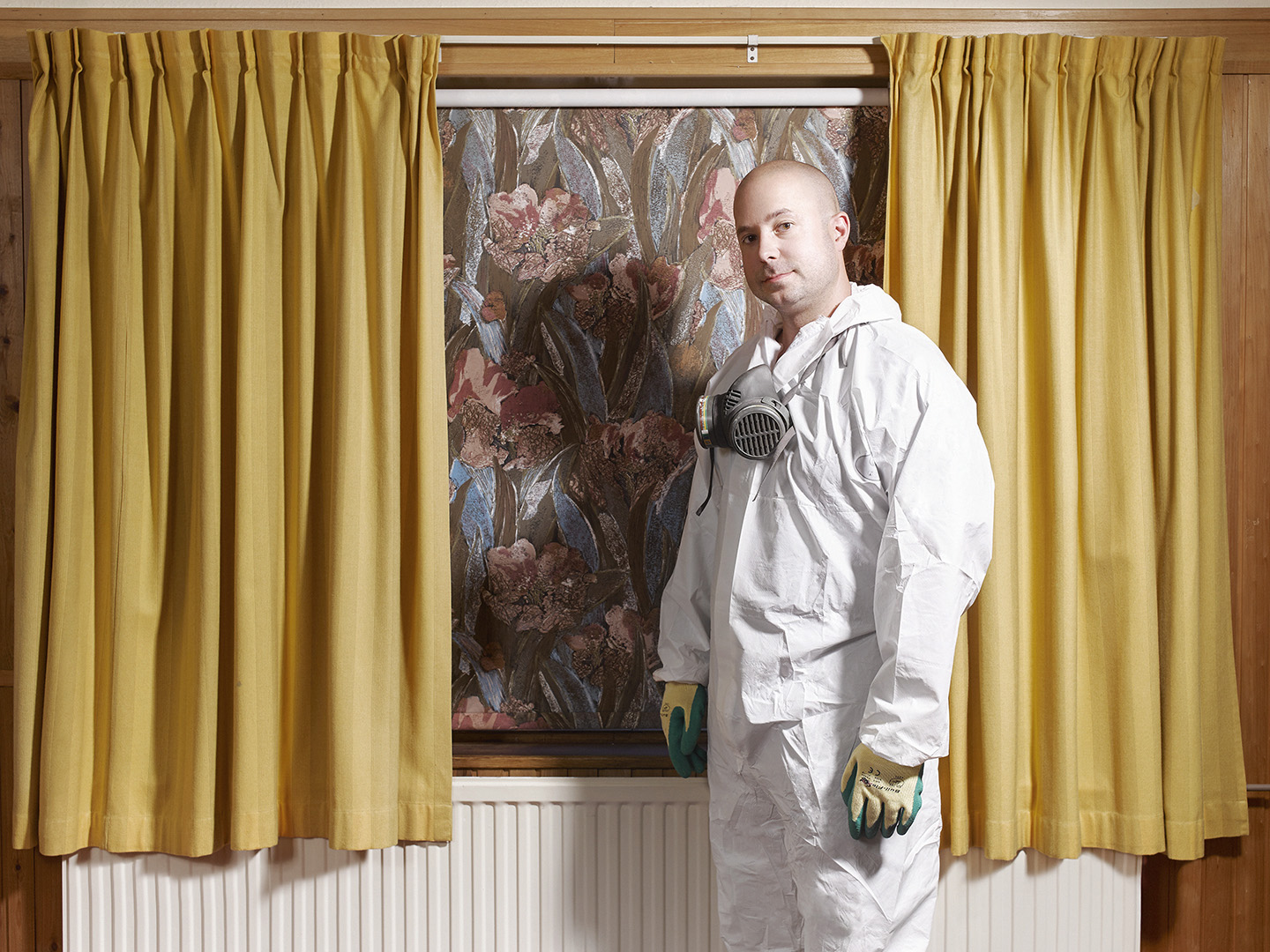 Man in a white hazmat suit and respirator stands indoors in front of floral wallpaper and yellow curtains, looking at the camera.