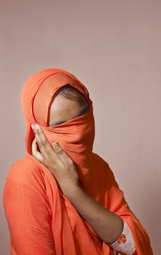 A portrait of an adult female in a bright orange robe covering her head and face, with only her eyes partially visible, against a peach background.