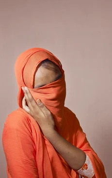 A portrait of an adult female in a bright orange robe covering her head and face, with only her eyes partially visible, against a peach background.