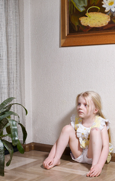 Blonde girl sits on floor, looking sideways near houseplant and painting of water lilies in home interior setting.