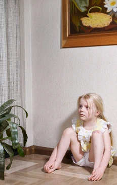 Blonde girl sits on floor, looking sideways near houseplant and painting of water lilies in home interior setting.