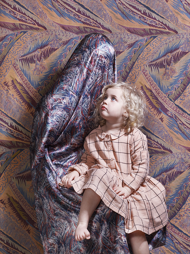 A toddler in a pink dress sits in front of a person draped in patterned fabric, both looking upward in a surreal, dreamlike setting.