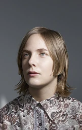 Portrait of a person with blonde hair wearing a patterned floral shirt against a grey background.