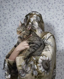 Person wrapped in a floral patterned fabric holding a tabby cat against a patterned grey wallpaper background.