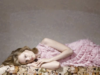 A woman in a pale pink fringed dress lying on a patterned sofa against a textured wall by Jouk Oosterhof.