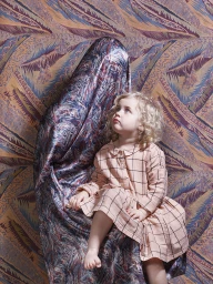 Young child with curly blonde hair wearing a pink plaid dress sitting on a person completely covered in patterned fabric