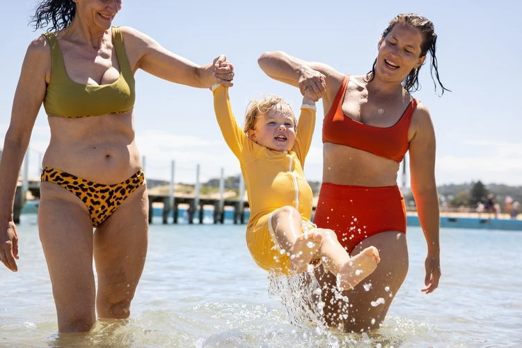 Two adult women hold a toddler in a yellow swimsuit above the water at the beach on a sunny day, creating splashes.