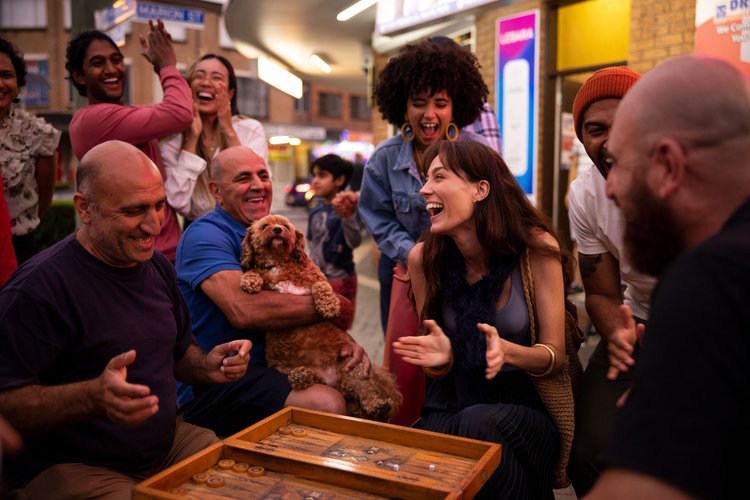 Group of people laughing outdoors on a city street, with a backgammon board, a brown dog, and city street signs in the background.