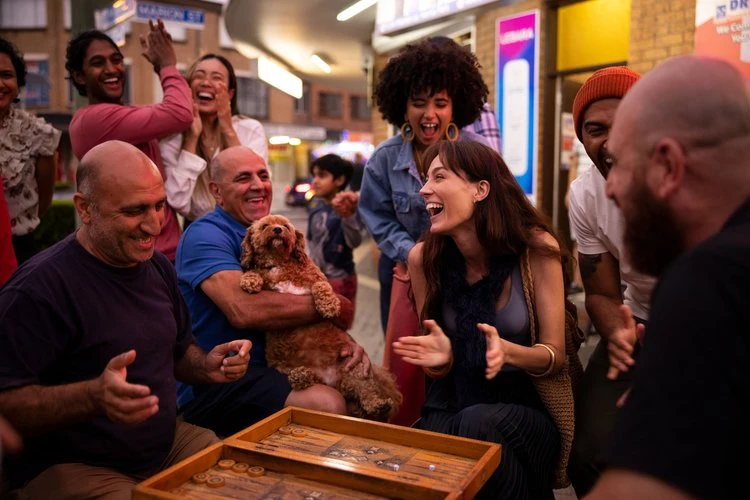 Group of people laughing outdoors on a city street, with a backgammon board, a brown dog, and city street signs in the background.