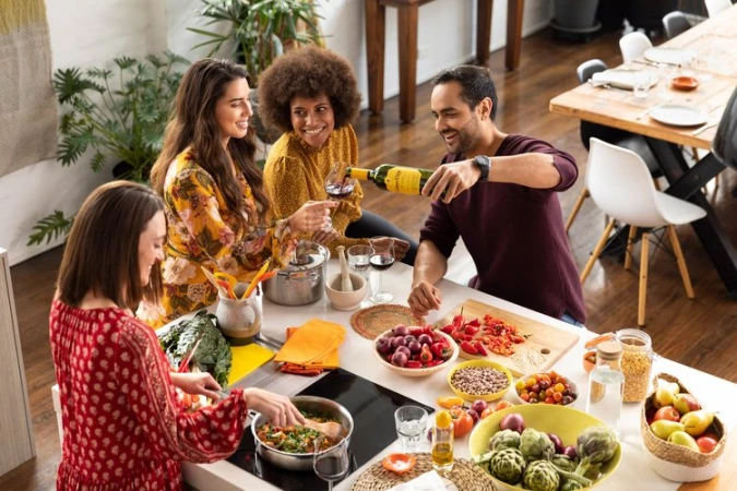 Four smiling adults preparing and serving a meal together at a kitchen island with fresh food and drinks in a brightly lit indoor space.