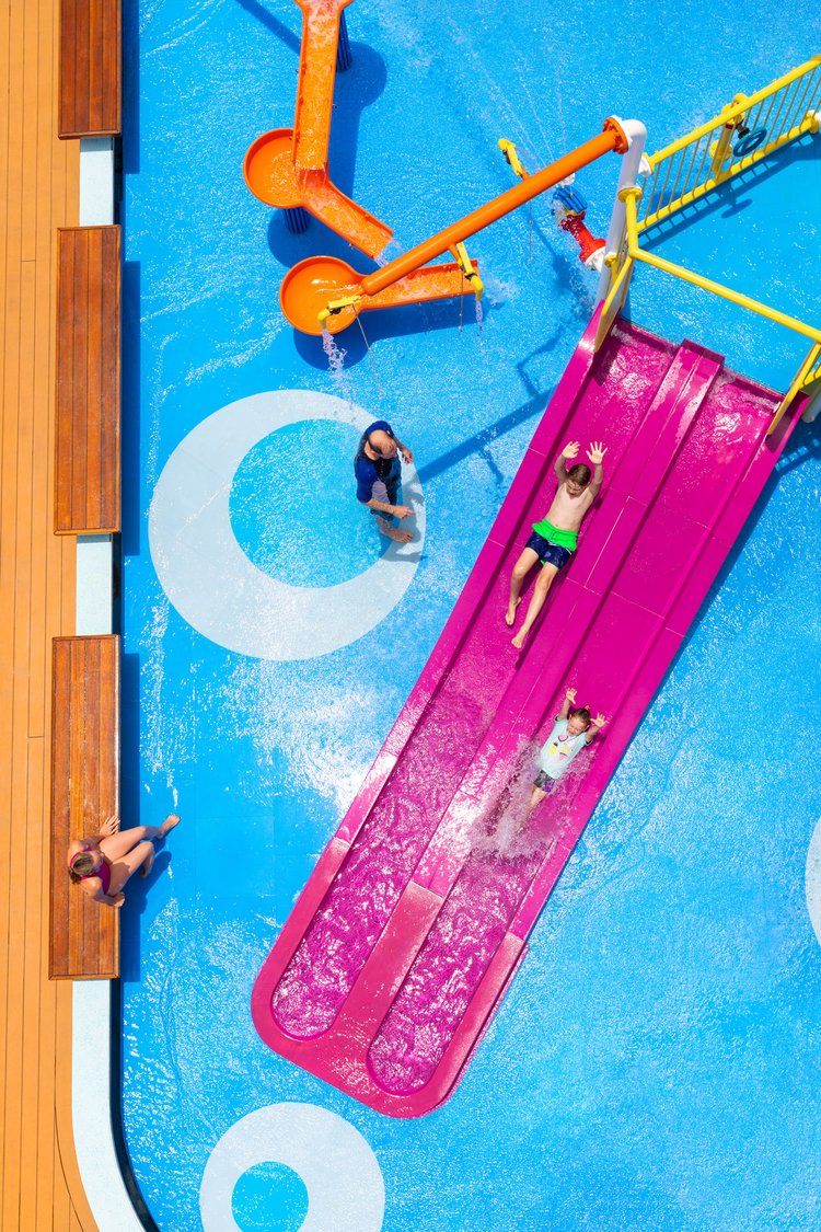 Aerial view of children on a water slide, with adults in the background, set against a blue and white pool deck with orange slides.