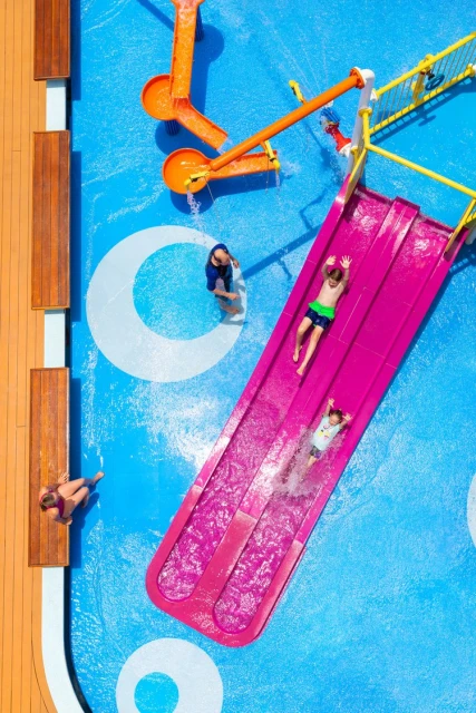 Aerial view of children on a water slide, with adults in the background, set against a blue and white pool deck with orange slides.