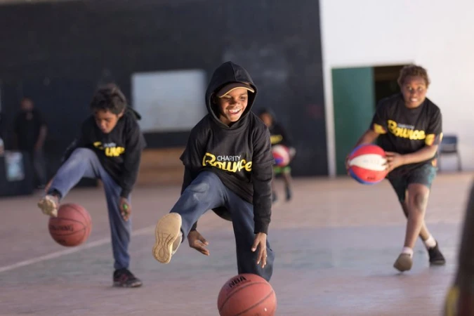 Three children in black shirts dribble basketballs on an indoor court, mid-motion, captured in a medium shot with diffused natural lighting.