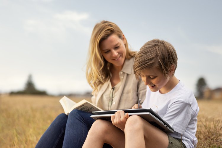 Adult female and child sit outdoors reading a book and drawing on a tablet; sunny daylight, blue sky, tall grass, neutral tones.