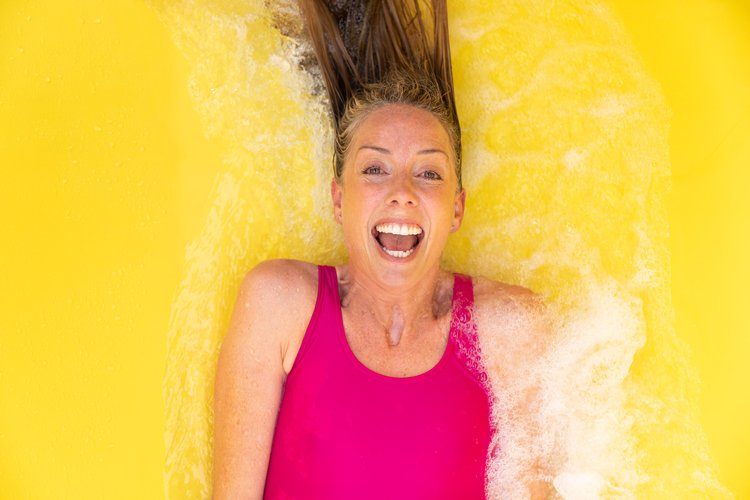 An adult woman with brown hair lies on her back in a bright yellow substance, expressing excitement and joy while wearing a pink swimsuit.
