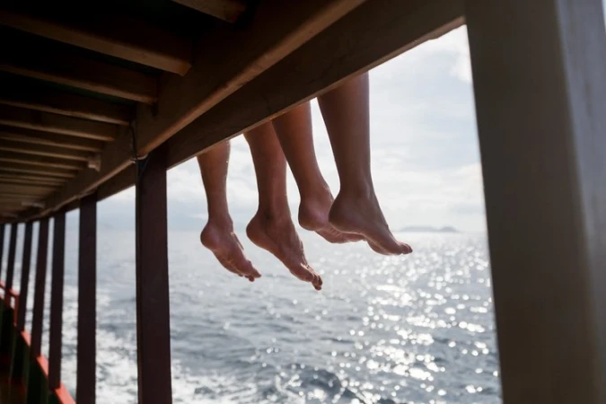 Close-up of bare legs dangling from a wooden boat deck overlooking the ocean water